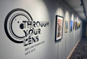 Framed art hanging on an illuminated wall labeled Through Your Lens Exhibit.