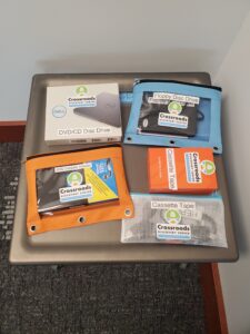 DVD disc drive, blue bag with floppy disc dirve, orange bag with VHS Cassette Adaptor, and orange box with cassette tapes on a tabletop.